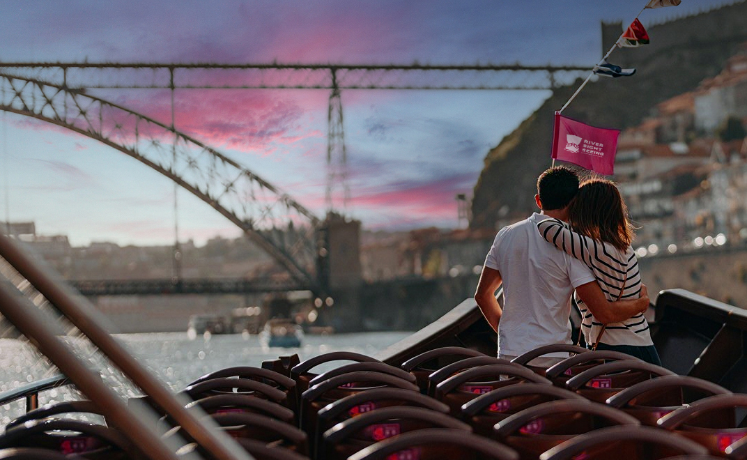 Casal abraçado na proa de um barco turístico, a olhar para a ponte sobre o rio Douro ao pôr do sol, com bandeirolas ao vento.