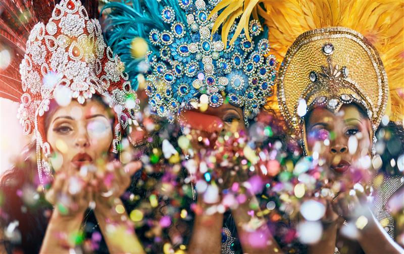 Three women in ornate carnival headdresses blow colorful confetti toward the camera, creating a festive bokeh effect.