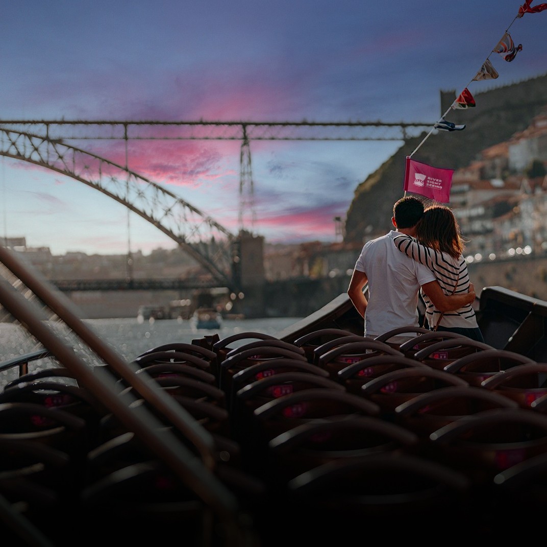 Casal abraçado na proa de um barco turístico, a olhar para a ponte sobre o rio Douro ao pôr do sol, com bandeirolas ao vento.