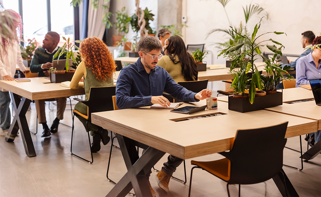 Employee working on a laptop at a shared table in a modern, plant-filled open office, with other coworkers in the background.