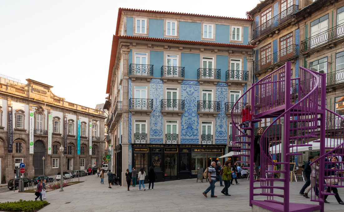 Praça no centro histórico do Porto com edifícios de fachadas tradicionais, incluindo azulejos azuis, e uma escultura/estrutura roxa com pessoas a circular.