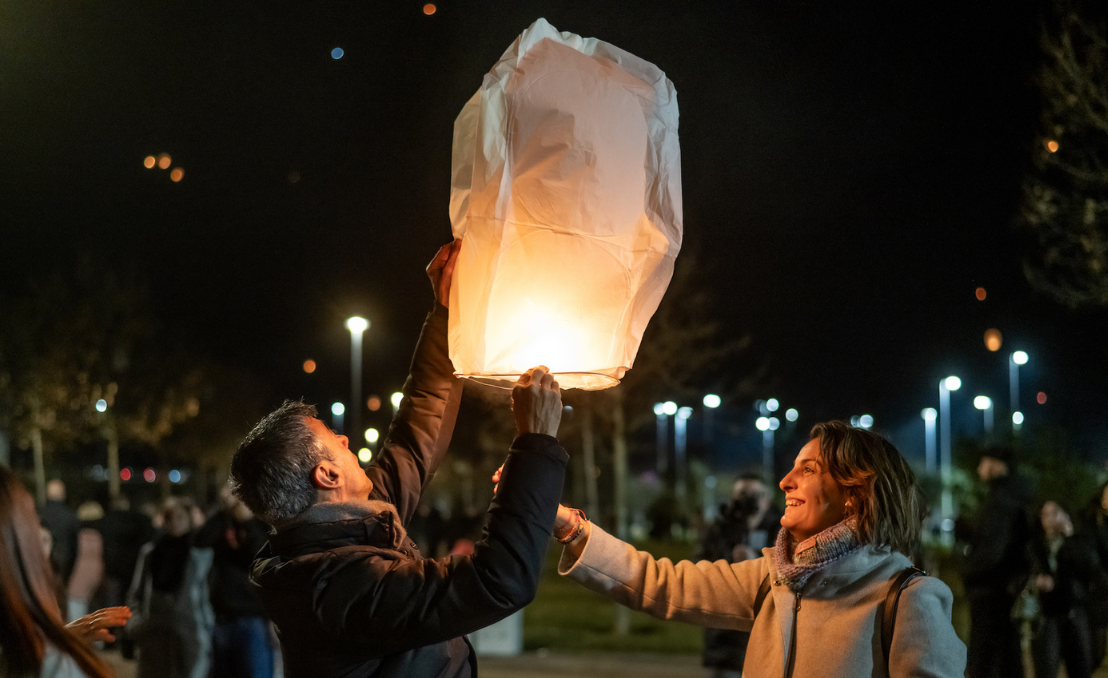 Duas pessoas a lançar um balão de São João iluminado, à noite, com luzes e multidão ao fundo.